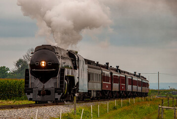 Fototapeta na wymiar A View of a Steam Passenger Train Approaching, Traveling Thru Rural America, Blowing Smoke on a Summer Day