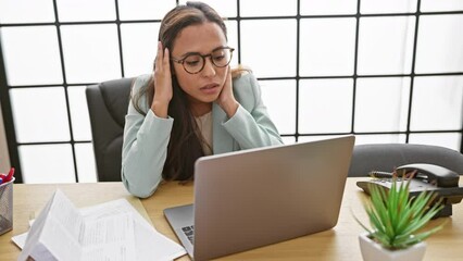 Overworked and stressed, the plight of a young, beautiful hispanic woman struggling with her laptop at her business workplace, juggling the pressures of her demanding job indoors.