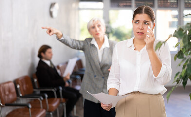 Upset young woman in business clothes with documents in her hands being criticized in office