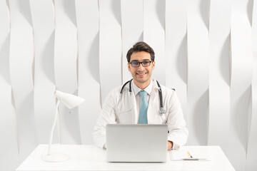 Young handsome physician in white medical gown smiling and working on laptop in his modern workplace