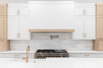 A kitchen detail with gold faucet and hardware, a marble subway tile backsplash, a large stove hood, and white and white oak cabinets. - obrazy, fototapety, plakaty