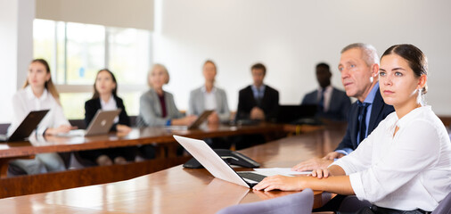 Side portrait of absorbed young white dark-haired businesswoman sitting with late-middle-aged white male colleague beside at conference table in boardroom, intently watching coworker's presentation
