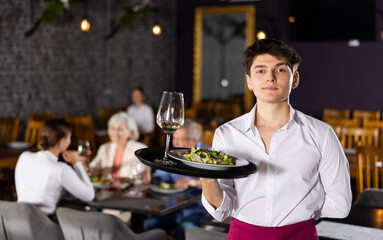 Young waiter guy is standing with order tray in spacious guest room in restaurant. Male restaurateur smiles and looks at camera. Cafe patrons eating in background. - obrazy, fototapety, plakaty