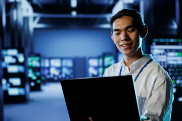 Smiling IT consultant in data center uses laptop to prevent system overload during peak traffic...