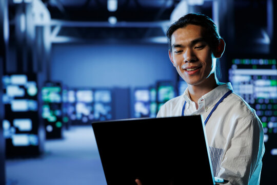 Smiling IT consultant in data center uses laptop to prevent system overload during peak traffic periods. Happy expert in server farm ensuring enough network bandwidth for smooth operations