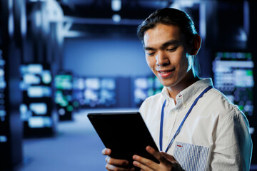Cheerful computer operator in data center uses tablet to prevent system overload during peak...