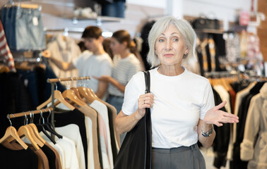 Pleased mature woman watching excitedly large stock of clothes in retail outlet
