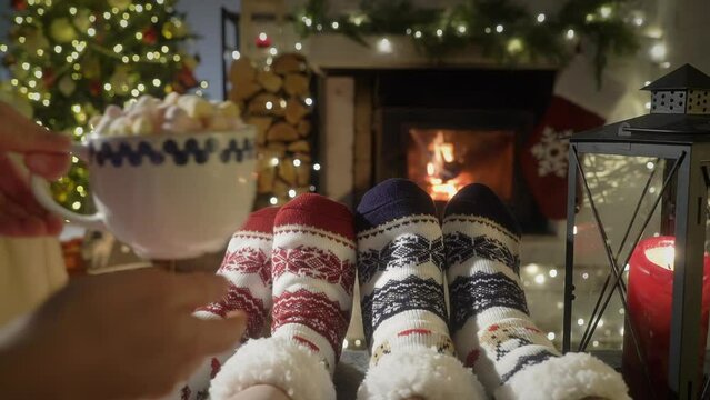 Couple enjoying by warm fireplace and warming up their feet in woolen socks with Christmas ornaments and Christmas tree and decorations background. Cozy Winter, Christmas and Family.