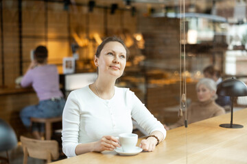 Dreamy smiling brown-haired woman sitting at table in cozy modern cafe with cup of aromatic coffee during breakfast