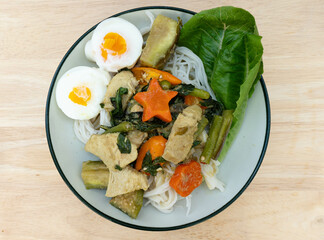 Thai rice noodles with Thai green curry with chicken, eggplant, basil, yellow chilli, vegetables and boiled eggs on a white dish and on a wooden background. Thai spicy food on the table