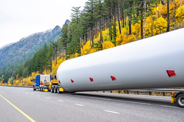 Blue big rig semi truck tractor transporting oversize load wind turbine pillar part on a semi trailer with a special additional trolley running on the autumn highway road with yellow mountain hills