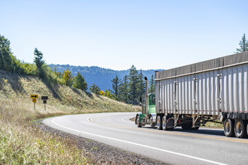 Classic green day cab big rig semi truck transporting cargo in bulk semi trailer moving on the winding mountain narrow road