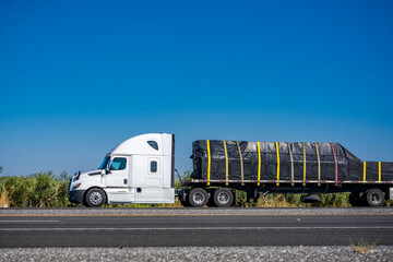 High cab white big rig semi truck transporting covered and fastened cargo on flat bed semi trailer...