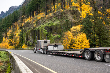 Gray classic powerful big rig semi truck with empty step deck semi trailer driving on the highway road with autumn yellow trees on the mountain ridge in Columbia River Gorge