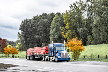 Low cab profile blue big rig bonnet semi truck tractor with covered cargo on loaded flat bed semi trailer standing on the street parking in green zone with autumn yellow trees at raining weather
