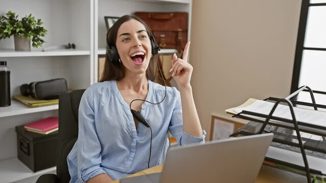 Young beautiful hispanic woman wearing headset smiling with an idea or question pointing finger up with happy face, number one at the office