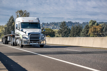 Low cab profile industrial big rig white semi truck transporting empty flat bed semi trailer running on the one way highway road