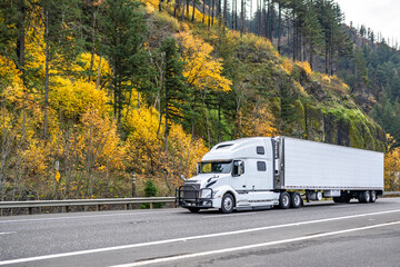 Pro industrial big rig white semi truck with grille guard transporting cargo in refrigerated semi trailer driving on the autumn road with yellow trees on the hill
