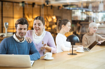 Positive European man and woman drinking and enjoying coffee while using laptop in cafe