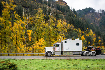 White big rig classic semi truck tractor with extended cab and step deck heavy duty semi trailer running on the autumn highway road