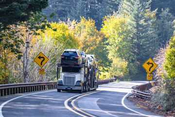 White classic car hauler big rig semi truck transporting cars on hydraulic semi trailer driving on...