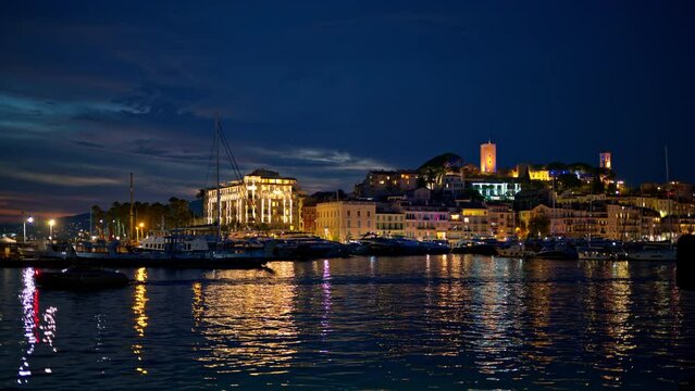 View of the sea port of Cannes with moored and floating yachts at night, France. Cityscape with Castre museum on a hill in the distance, illumination