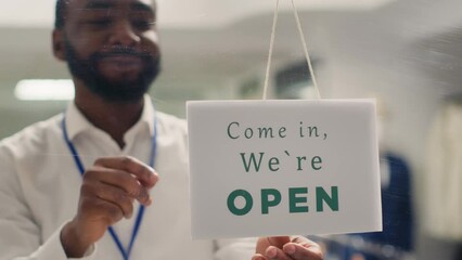 African american man working in clothing store opening in the morning, waiting for customers. Happy BIPOC retail clerk turning closed message sign on fashion boutique door in mall
