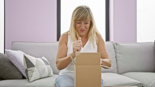 Joyful middle age blonde woman delightfully unpacking a cardboard box at her beautiful home, sitting confidently on the living room sofa, smiling wide, indoors, shipping parcel delivered