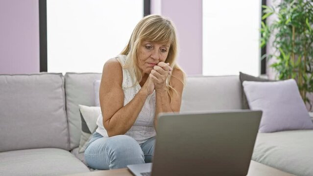 In her cozy home, a serious and focused middle age blonde woman is sitting on the sofa, totally absorbed, using her laptop in the living room's welcoming interior.