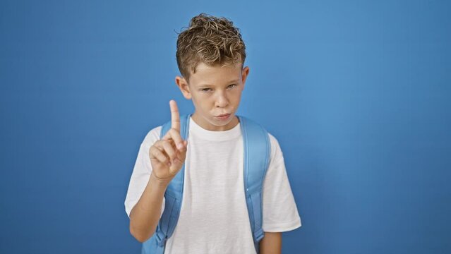 Adorable blond boy student, a smart little kid standing, serious face expressing negation, saying no with his finger. an isolated blue wall cutout background accentuates his academic bag.
