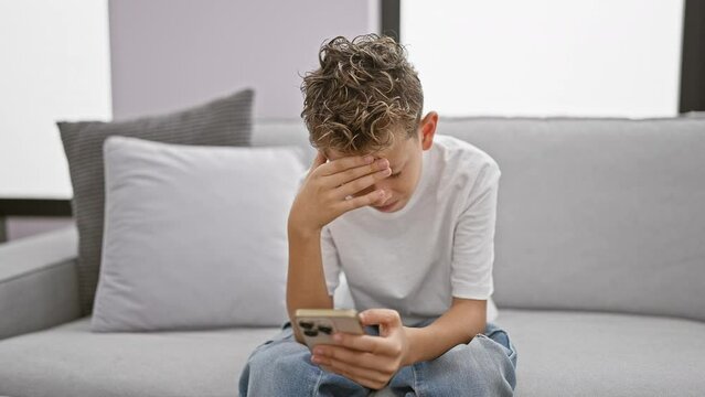 Adorable blond boy, stressed and upset, sitting on sofa at home using smartphone. worried child typing a message, showing serious expression amidst technology stress.