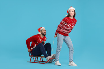 Young couple in Christmas sweaters. Woman pulling her man in sled on light blue background