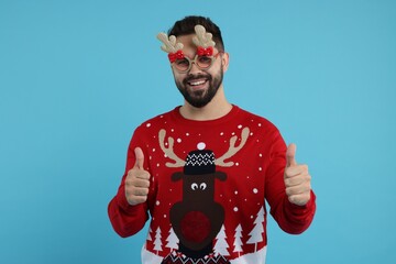 Happy young man in Christmas sweater and funny glasses showing thumbs up on light blue background