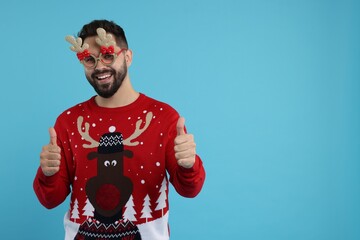Happy young man in Christmas sweater and funny glasses showing thumbs up on light blue background. Space for text
