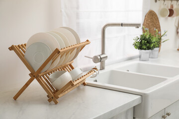 Drying rack with clean dishes on light marble countertop near sink in kitchen - obrazy, fototapety, plakaty