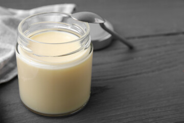 Open jar with condensed milk and spoon on dark grey wooden table, closeup. Space for text
