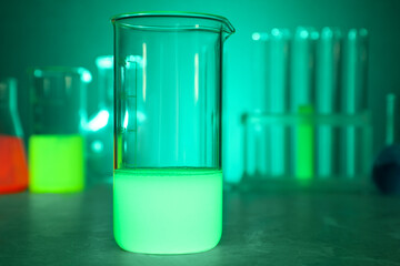Laboratory beaker with luminous liquid on table against blurred background, closeup
