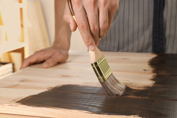 Man with brush applying wood stain onto wooden surface indoors, closeup
