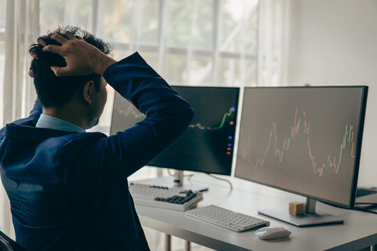 Man groans with hands on computer head with graph of unsuccessful business on monitor screen with cryptocurrency graph that tracks cryptocurrency prices.