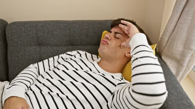 Stressed young hispanic man alone, grappling with anxiety, lies in despair on his living room sofa at home, a portrait of frustration and sadness