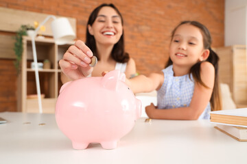 Little girl with her mother putting coin into piggy bank at home, closeup