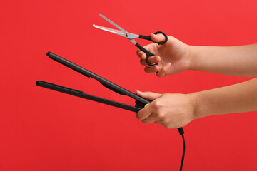 Female hands with straightening iron and scissors on red background.