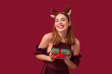Beautiful young woman in reindeer horns with Christmas gift box on red background