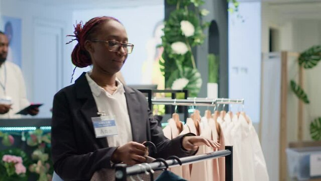 Portrait of woman restocking elegant selection of shirts on hangers in high end fashion boutique. Happy worker arriving with trendy, stylish garments from new collection in clothing shop