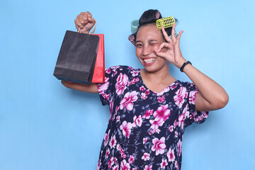 Happy Asian shopaholic housewife showing shopping bags and ok fingers while wearing hair curlers...
