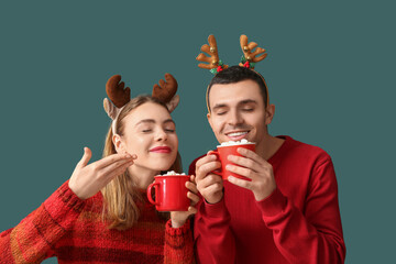 Beautiful young couple in reindeer horns holding cups of coffee with marshmallows on green background