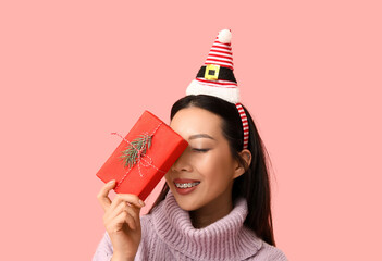 Young Asian woman in Elf hat headband with Christmas gift box on pink background