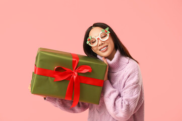 Young Asian woman in decorative eyeglasses with Christmas gift box on pink background