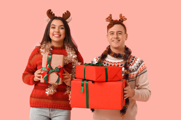 Young couple in reindeer horns with Christmas tinsel and gift boxes on pink background