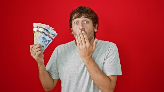 Shocked young blond man covering mouth in surprise and fear, holding canadian dollar banknotes over red isolated background - afraid of his own money mistake in silence
